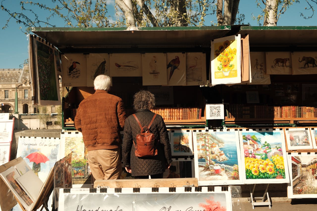 A couple explores an art stall in Paris under the sun, displaying vibrant paintings.
